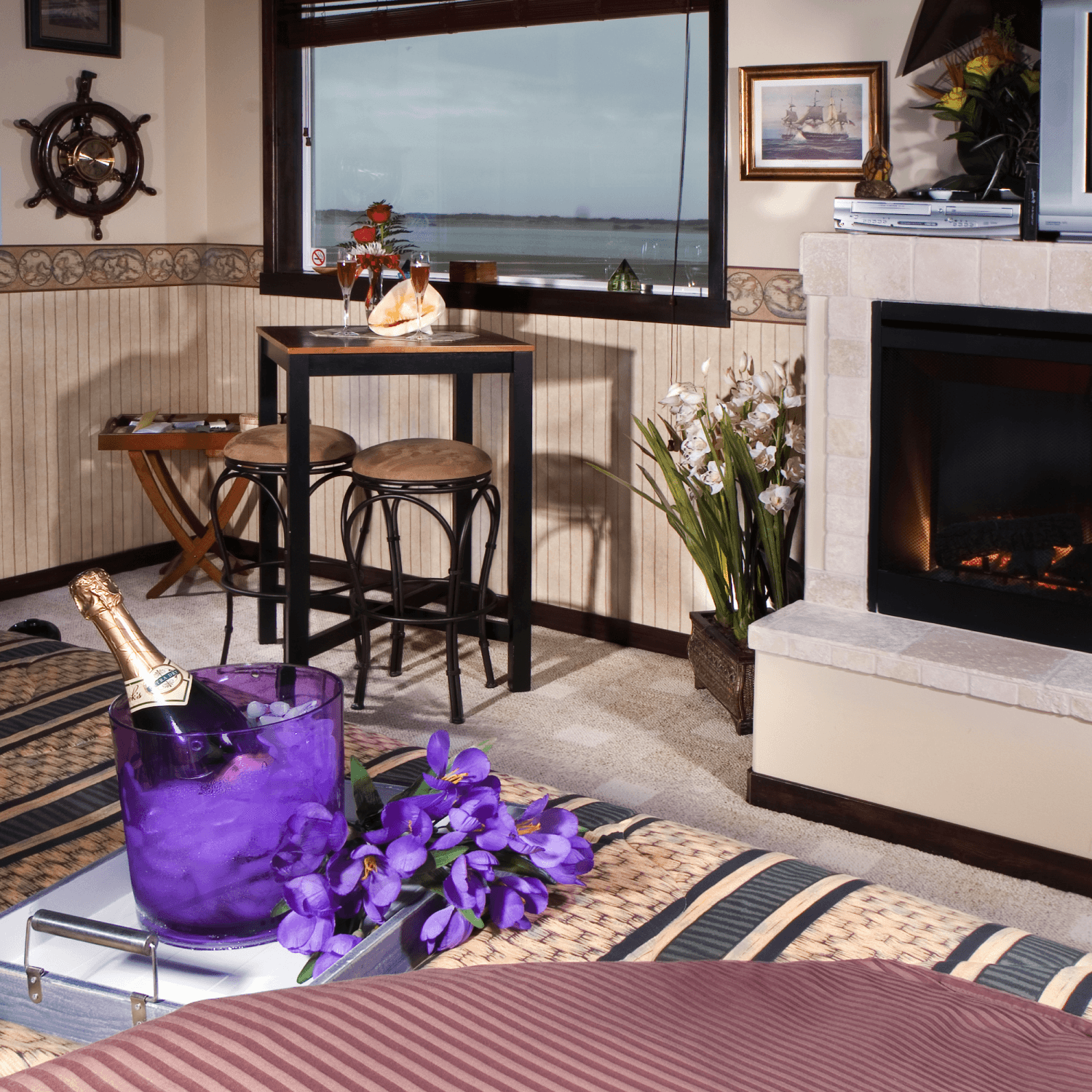 Cozy interior of a stylish room featuring a fireplace, a table with drinks, and decorative flowers, with windows showcasing a scenic view.