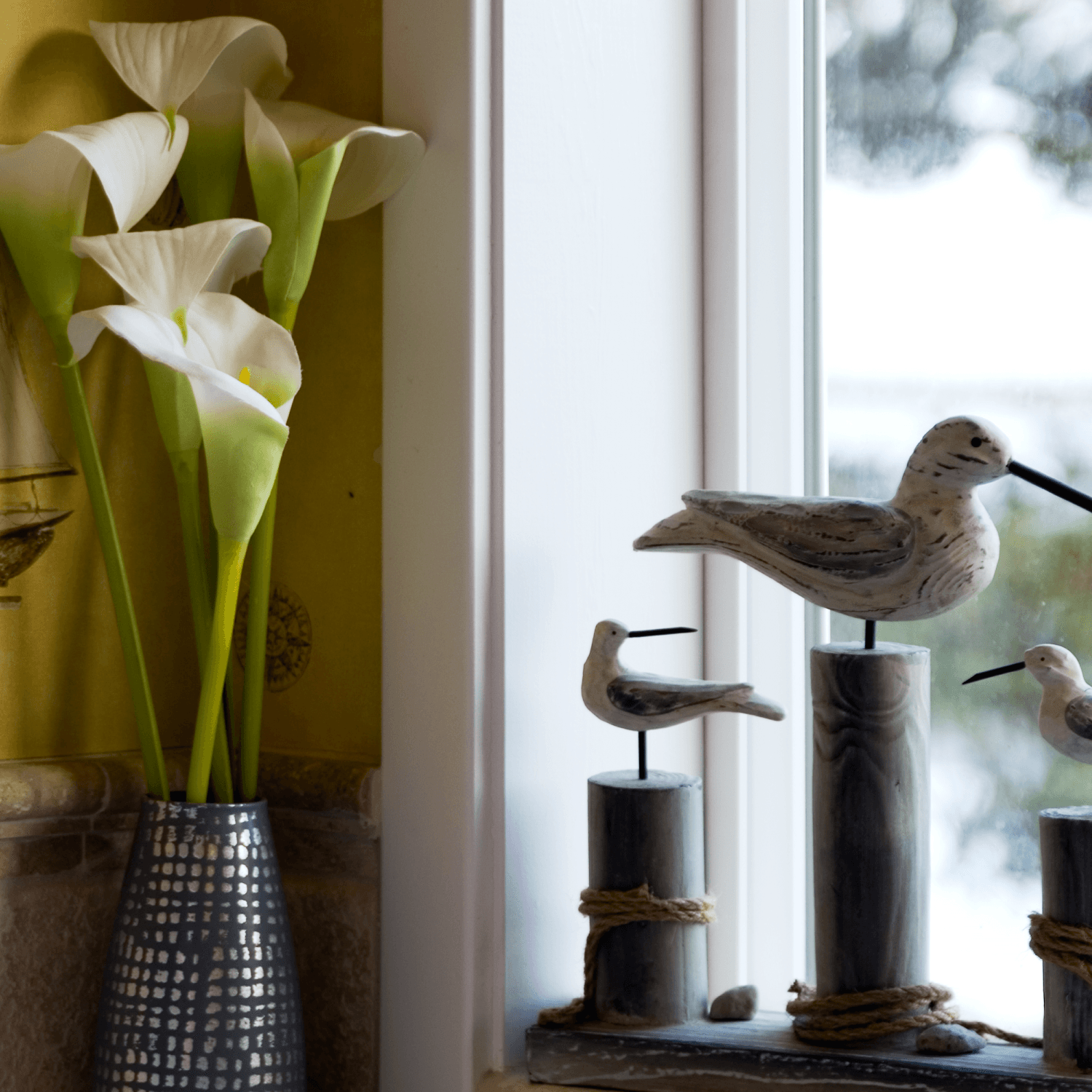 A vase of white calla lilies beside decorative wooden birds on a windowsill.